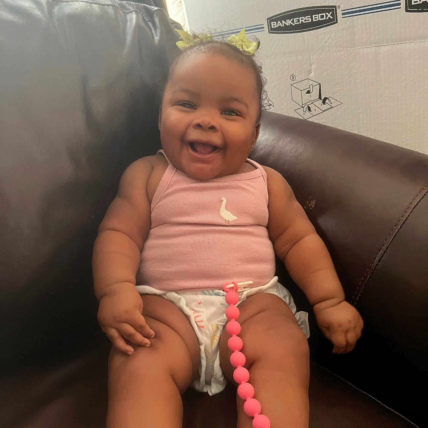 Jireh is registered to the contest to win money with this photo: baby, child, smiling, sitting, pink_clothing, hair_bows, diaper, pacifier, beaded_chain, leather_chair, cardboard_box, happy, cute, indoors, person, infant, plump_arms, plump_legs, casual, portrait