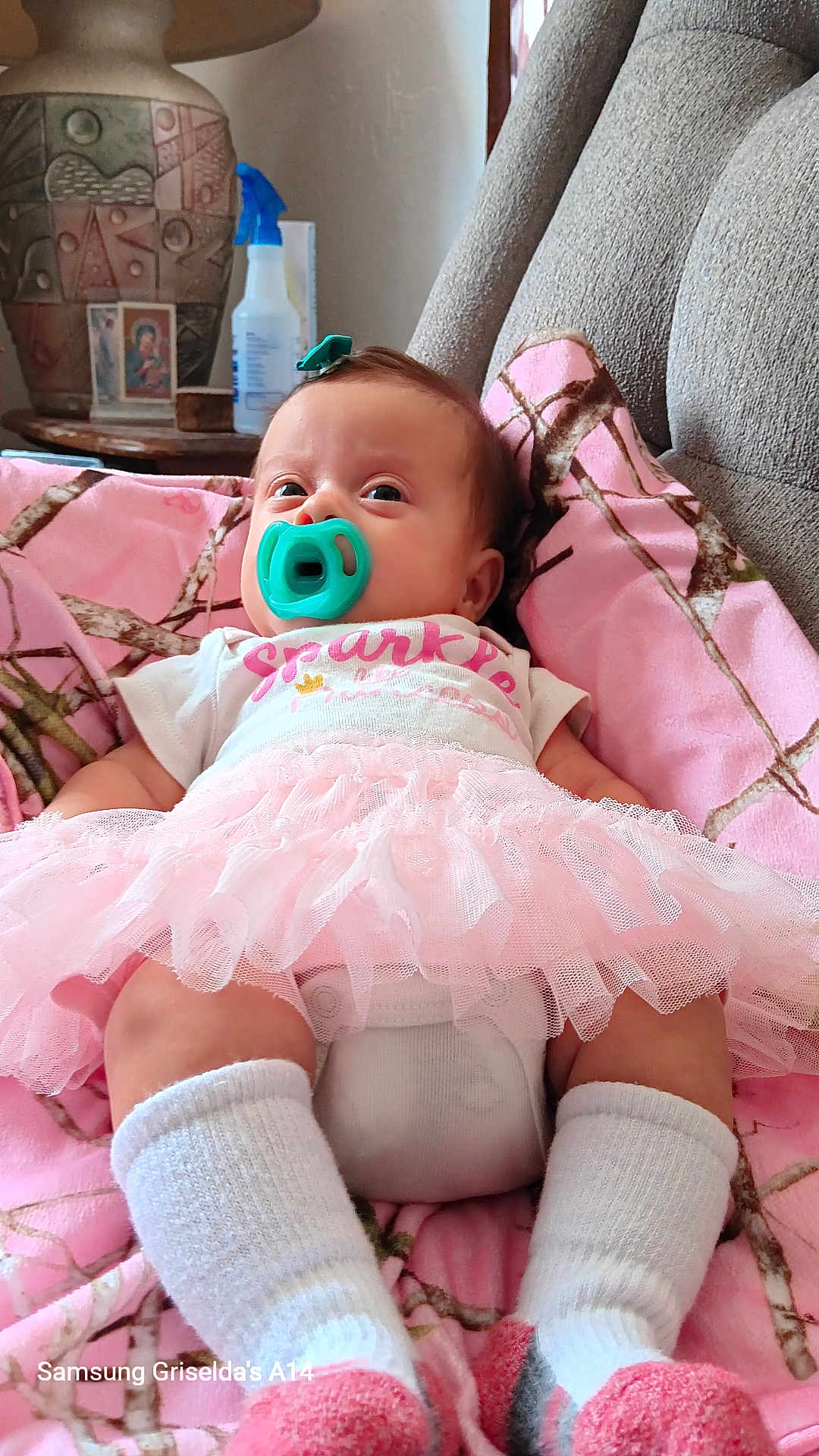 Naomi is registered to the contest to win money with this photo: baby, pacifier, tutu, pink_blanket, white_socks, hair_clip, armchair, vase, table, indoor, child, cute, infant, clothing, comfort, portrait, relaxing, home, soft, cozy