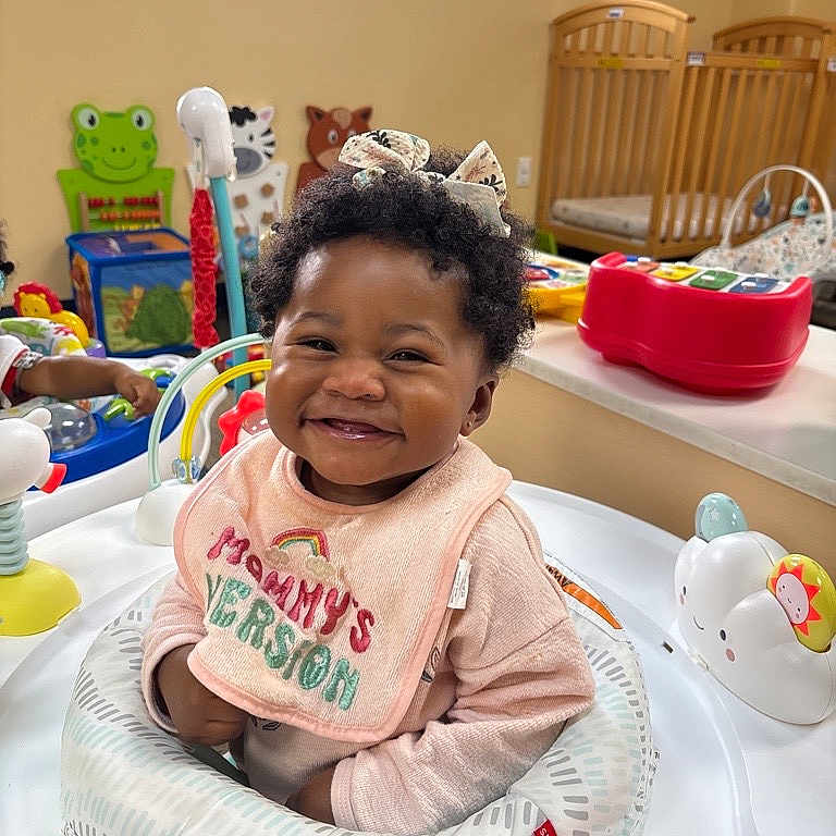 Alora is registered to the contest to win money with this photo: activity_center, baby, bib, bow, child, clothing, colorful, crib, curly_hair, cute, early_childhood, happy, indoor, infant, nursery, person, playroom, seat, smiling, toy