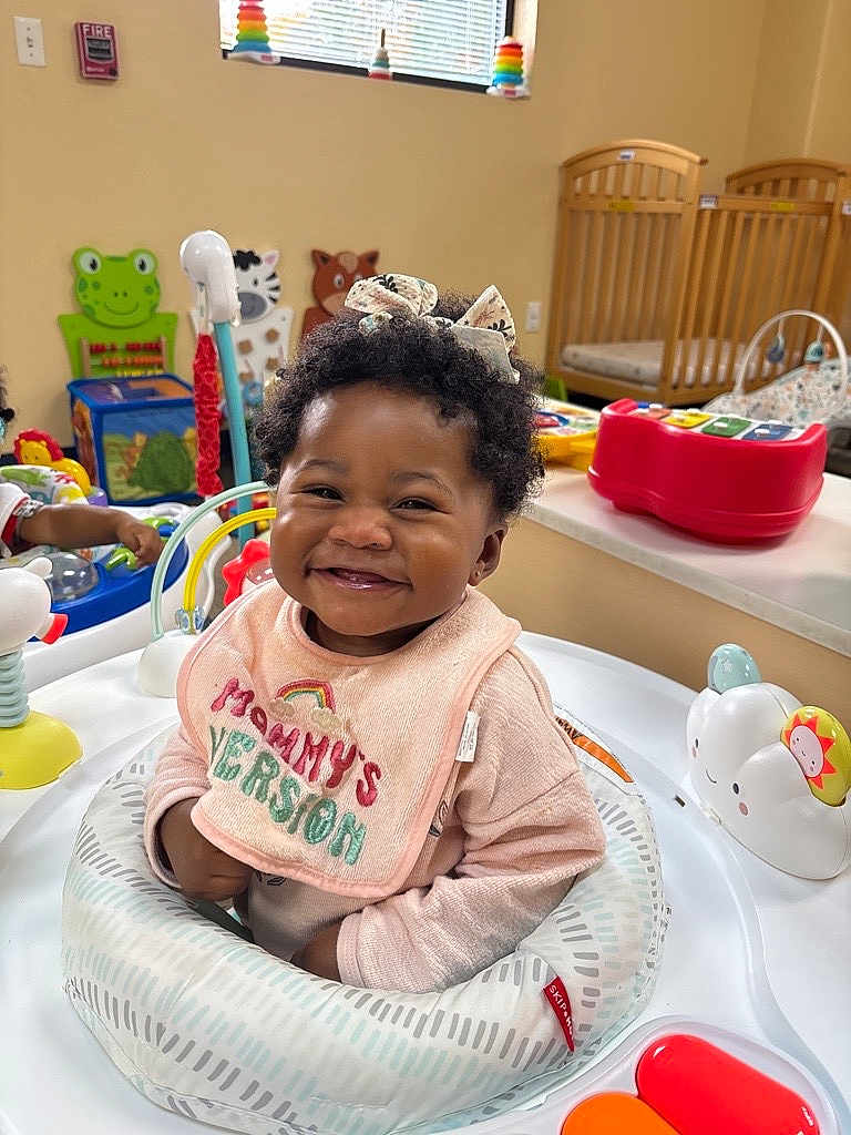 Alora is registered to the contest to win money with this photo: baby, smiling, child, toy, activity_center, bow, curly_hair, nursery, crib, colorful, playroom, infant, clothing, bib, happy, indoor, person, cute, seat, early_childhood