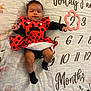 baby, baby_clothing, black_socks, celebration, child, cute, fabric, growth_tracker, indoors, infant, ladybug_costume, lying_down, milestone_blanket, months_marker, pattern, person, polka_dots, portrait, red_dress, smiling