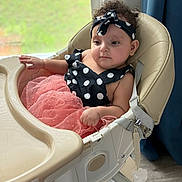 Serafina is registered to the contest to win money with this photo: baby, high_chair, polka_dot, pink_skirt, headband, curly_hair, indoor, window, natural_light, child, cute, infant, seated, clothing, portrait, young_child, furniture, looking_away, skin, earrings