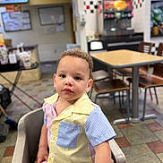 Simon is registered to the contest to win money with this photo: toddler, child, high_chair, curly_hair, striped_shirt, indoor, restaurant, table, chair, floor_tiles, face, person, casual_clothing, portrait, seated, young_child, curious_expression, light_skin, short_sleeves, dining_area