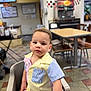 toddler, child, high_chair, curly_hair, striped_shirt, indoor, restaurant, table, chair, floor_tiles, face, person, casual_clothing, portrait, seated, young_child, curious_expression, light_skin, short_sleeves, dining_area