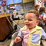 Simon is registered to the contest to win money with this photo: toddler, child, smiling, chair, indoor, restaurant, food, snack, multicolored_shirt, curly_hair, happy, person, casual, background_people, menu, table, seat, light, portrait, cute