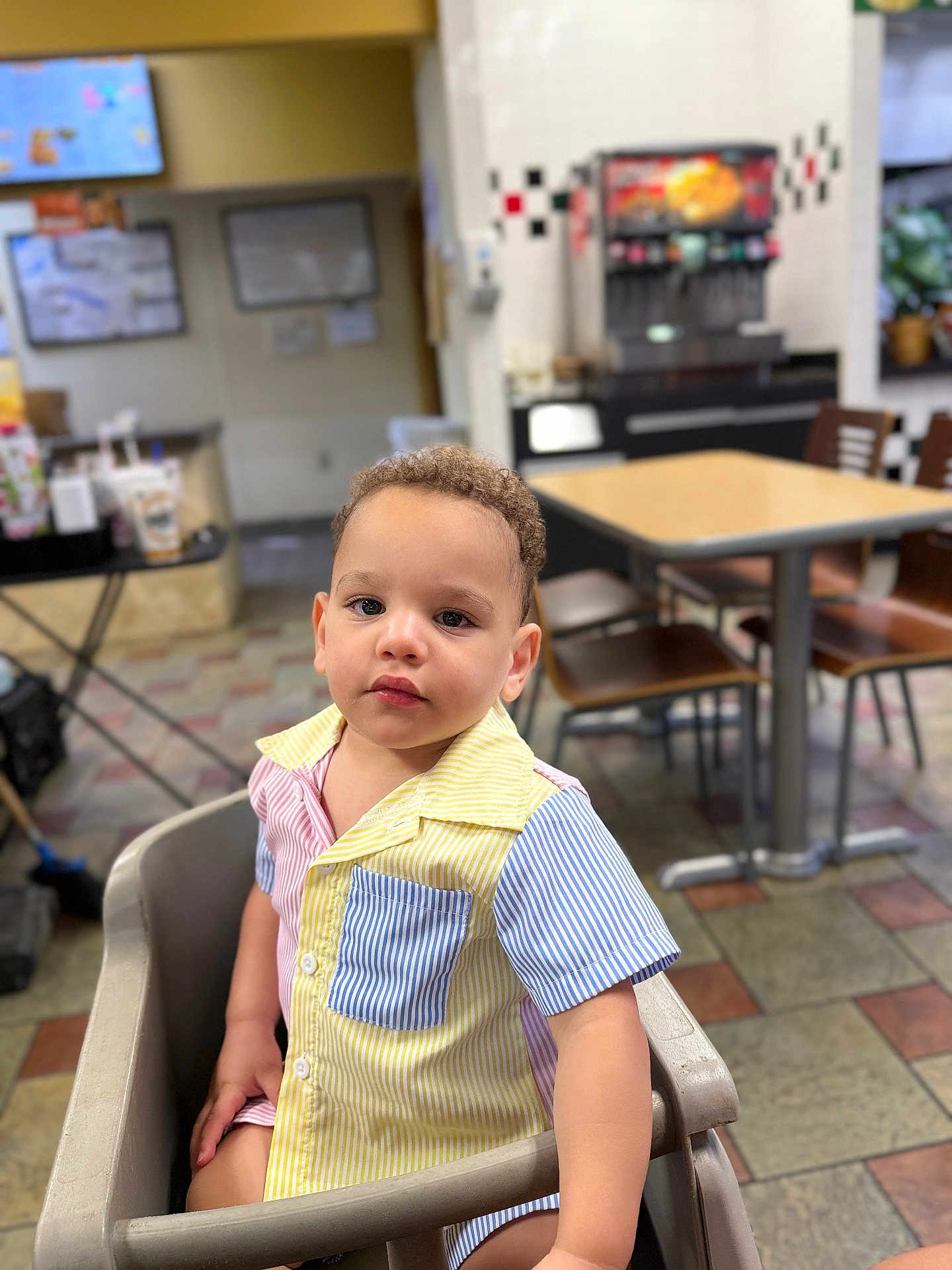 Simon joined the competition — help win amazing prizes! toddler, child, high_chair, curly_hair, striped_shirt, indoor, restaurant, table, chair, floor_tiles, face, person, casual_clothing, portrait, seated, young_child, curious_expression, light_skin, short_sleeves, dining_area