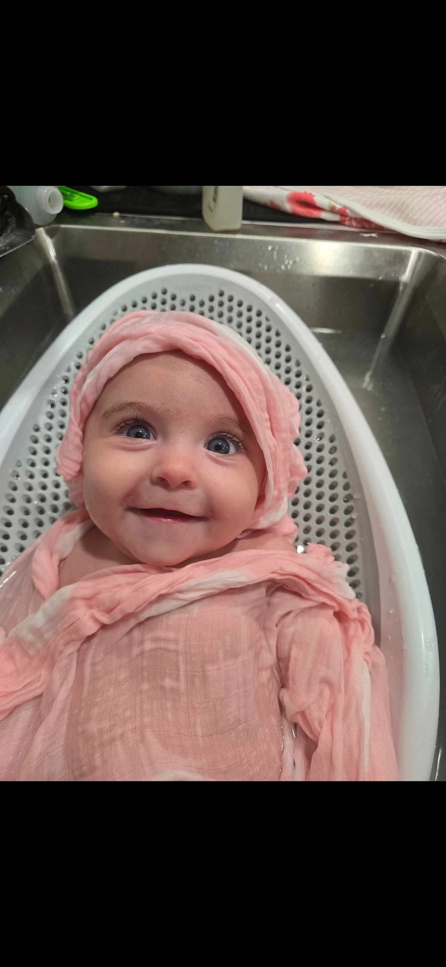 Letty is registered to the contest to win money with this photo: baby, infant, smile, pink_cloth, bath, white_bath_seat, sink, water, blue_eyes, wrapped, happy, cute, child, face, indoor, towel, kitchen_sink, person, head_covering, closeup