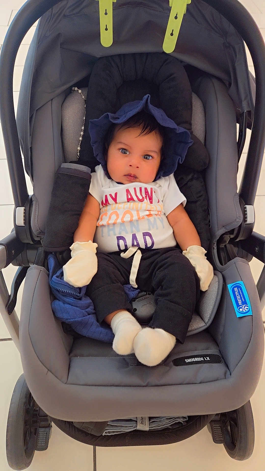 Zayn is registered to the contest to win money with this photo: baby, stroller, infant, mittens, blue_bonnet, white_socks, black_pants, colorful_shirt, face, child, tiled_floor, wheels, car_seat, cute, seated, indoors, expression, clothing, person, portrait