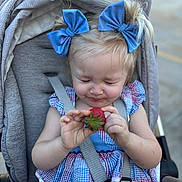 Hazel is registered to the contest to win money with this photo: toddler, child, stroller, blue_bows, strawberry, dress, checkered, outdoor, seatbelt, cute, funny_expression, hair_bows, baby, person, summer, food, fruit, small_child, portrait, daylight