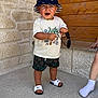 toddler, child, smiling, hat, bucket_hat, shorts, sandals, sunglasses, stone_wall, wood_panel, outdoor, standing, happy, summer, casual_clothing, footwear, person, cute, daylight, playful