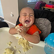 Uriyah joined the competition — help win amazing prizes! baby, child, high_chair, food, mashed_food, messy, happy, laughing, orange_shirt, indoor, table, sippy_cup, feeding, cute, fun, face, hands, window, light, smile