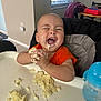 baby, child, high_chair, food, mashed_food, messy, happy, laughing, orange_shirt, indoor, table, sippy_cup, feeding, cute, fun, face, hands, window, light, smile