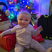 Lillian joined the competition — help win amazing prizes! baby, child, christmas_tree, present, gift_box, pajamas, holiday, indoor, smile, festive, lights, carpet, holiday_decor, cute, sitting, cheerful, celebration, colorful, winter, joy