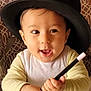 baby, child, hat, magic_wand, smiling, happy, indoor, table, cushion, patterned, person, face, hand, clothing, sitting, cute, playful, young_child, headwear, portrait