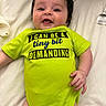 baby, infant, smiling, clothing, green_onesie, text_on_clothing, lying_down, bed, white_sheet, hand, face, child, cute, happy, portrait, skin, infant_clothing, young_child, indoors, person