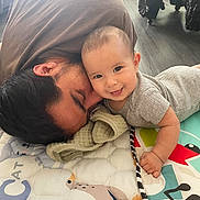 Julian is registered to the contest to win money with this photo: baby, adult, child, smiling, play_mat, floor, towel, cuddling, indoor, happy, person, head, face, clothing, wooden_floor, toy, colorful, cute, family, bonding