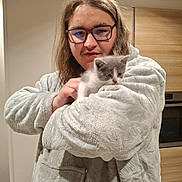 Yuko participe au concours pour gagner de l'argent avec cette photo : person, glasses, kitten, cat, indoor, robe, furry_clothing, holding, kitchen, wooden_cabinet, appliance, curious, pet, cute, young_adult, casual, home, portrait, animal, cozy