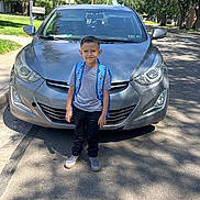 Bradley joined the competition — help win amazing prizes! child, boy, backpack, car, street, sidewalk, trees, shadows, sunny, casual_clothing, sneakers, residential_area, outdoor, daytime, vehicle, gray_car, curb, greenery, person, smile