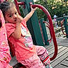 child, toddler, playground, playground_equipment, outdoors, park, sitting, handrail, red_bars, green_panel, wooden_deck, trees, nature, daylight, pink_shirt, hello_kitty, patterned_pants, sneakers, smiling, portrait