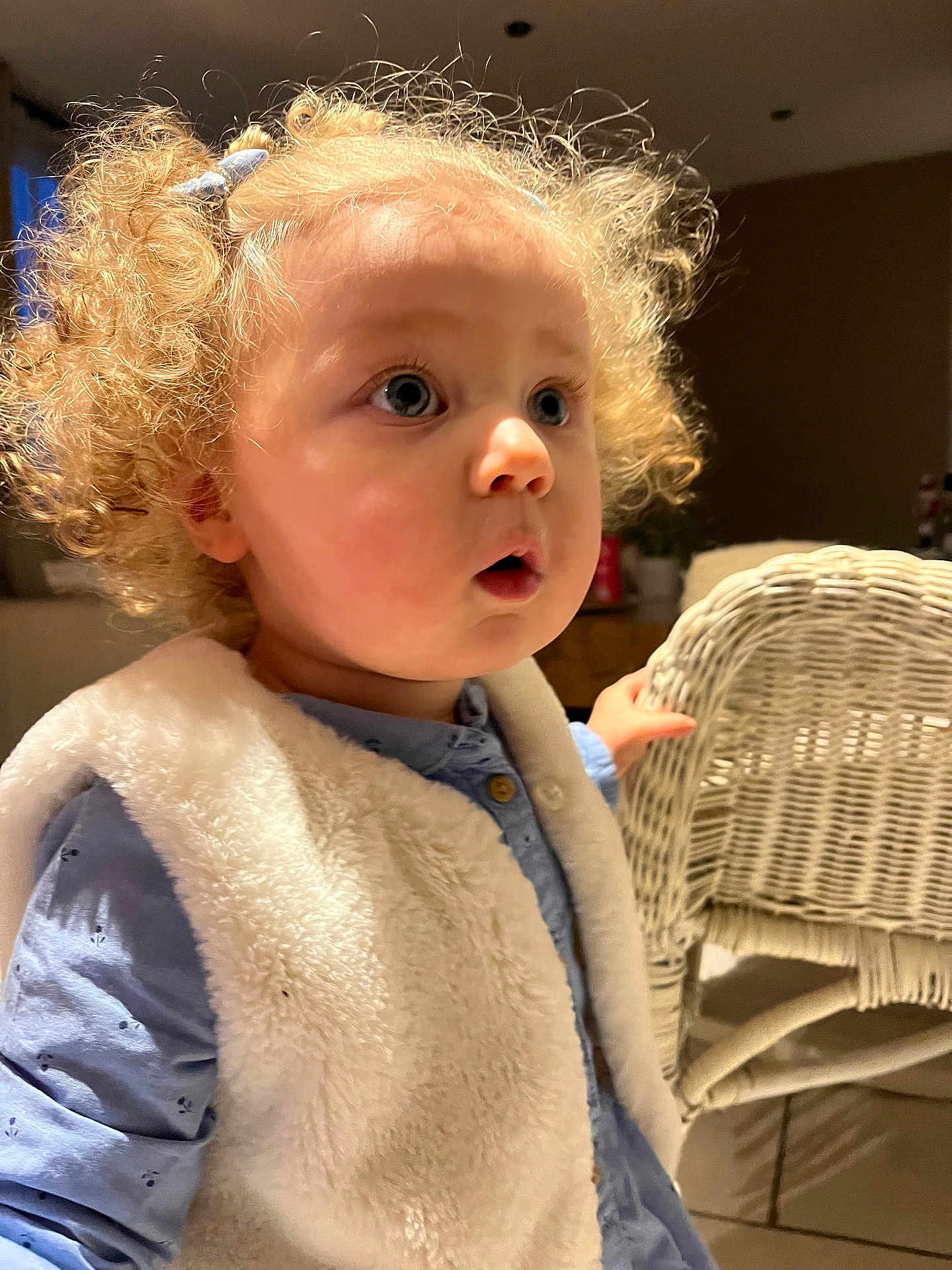 Alba a rejoint le concours — aidez-le/la à gagner de superbes lots ! toddler, child, curly_hair, blonde_hair, blue_eyes, vest, blue_shirt, indoor, warm_light, wicker_chair, hand, curious_expression, person, portrait, cute, soft_texture, clothing, hairclip, face, furniture