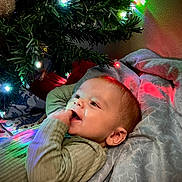 Cyrus is registered to the contest to win money with this photo: baby, child, christmas_tree, lights, blanket, sweater, holiday, indoors, smiling, face, cozy, colorful_lights, soft_texture, infant, cute, relaxed, decorations, festive, portrait, happy