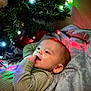 baby, child, christmas_tree, lights, blanket, sweater, holiday, indoors, smiling, face, cozy, colorful_lights, soft_texture, infant, cute, relaxed, decorations, festive, portrait, happy