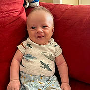 Cyrus is registered to the contest to win money with this photo: baby, infant, smiling, sitting, red_couch, dinosaur_pajamas, sea_creature_pajamas, indoors, window_light, happy, cute, child, person, clothing, furniture, portrait, soft_light, cozy, home, relaxed