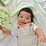 baby, smiling, infant, flower, basket, white_flower, blanket, checkered_outfit, cute, lying_down, happy, portrait, child, soft_lighting, indoors, small_hand, buttoned_overall, decor, closeup, young_child