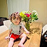 child, toddler, curly_hair, portrait, sitting, dining_table, flowers, bouquet, indoors, home, chair, wooden_table, red_shorts, gray_shirt, bare_feet, looking_at_camera, colorful, emoji, furniture, family_photo