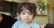 Ayhan Sharifi is registered to the contest to win money with this photo: toddler, child, curly_hair, portrait, indoors, home, kitchen, living_room, face, eyes, neutral_expression, gray_shirt, clothing, sitting, adult, arm, furniture, plant, apartment, photography