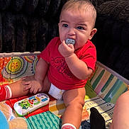 Kazimir is registered to the contest to win money with this photo: baby, infant, child, red_shirt, diaper, red_socks, toy_remote, plush_toy, play_mat, chewing, indoor, couch, colorful, cute, sitting, person, young_child, face, hand, foot