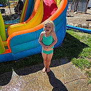 Oakley is registered to the contest to win money with this photo: child, swimsuit, inflatable_slide, outdoor, sunny, barefoot, grass, concrete, shadow, tree, people, fence, water, playground, summer, recreation, fun, daytime, person, leisure
