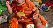 Zito is registered to the contest to win money with this photo: baby, child, infant, orange_onesie, sun_design, cushioned_seat, living_room, carpet, couch, blanket, baby_rocker, foot, hand, smile, face, indoors, window_blinds, home, comfort, cute