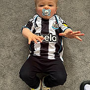 Brooklyn is registered to the contest to win money with this photo: baby, black_and_white_jersey, carpet, casual_wear, child, clothing, cute, expression, floor, footwear, indoor, lying_down, pacifier, person, reaching_hands, sneakers, soccer_jersey, sportswear, toddler, young_child