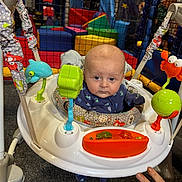 Finley Robinson is registered to the contest to win money with this photo: baby, infant, jumper, toys, indoor_play_area, colorful, hand, rings, carpet, plastic, seat, child, face, clothing, blue_outfit, play_equipment, expression, looking_at_camera, netting, soft_toys