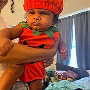 Christian joined the competition — help win amazing prizes! baby, child, infant, pumpkin_costume, orange_hat, costume, portrait, being_held, adult, face, curly_hair, indoor, bedroom, curtains, laundry, clothing, serious_expression, sitting, family, photography
