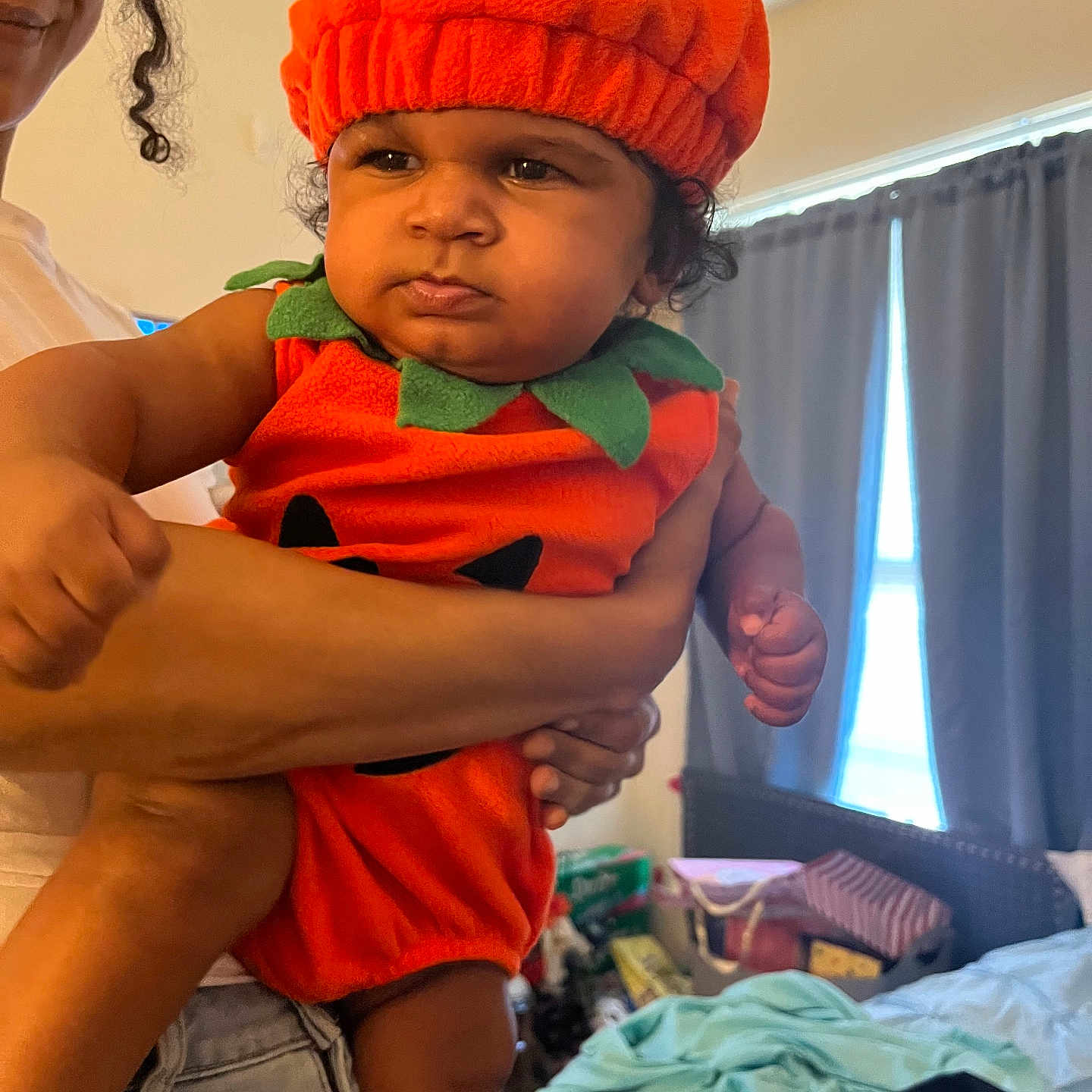 Christian joined the competition — help win amazing prizes! baby, child, infant, pumpkin_costume, orange_hat, costume, portrait, being_held, adult, face, curly_hair, indoor, bedroom, curtains, laundry, clothing, serious_expression, sitting, family, photography