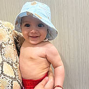 Aslan is registered to the contest to win money with this photo: baby, child, smiling, hat, blue_hat, bear_print, red_shorts, bracelet, hand, snake_pattern, person, indoor, portrait, cute, skin, face, happy, holding, background, wood