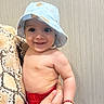 baby, child, smiling, hat, blue_hat, bear_print, red_shorts, bracelet, hand, snake_pattern, person, indoor, portrait, cute, skin, face, happy, holding, background, wood
