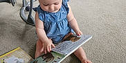 Brooklynn is registered to the contest to win money with this photo: baby, child, headband, reading, book, carpet, floor, indoor, childrens_book, curly_hair, sitting, casual_clothing, blue_dress, toys, learning, cute, infant, early_learning, playtime, education