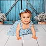 baby, child, blue_dress, curly_hair, crawling, floor, blue_eyes, bow, flowers, basket, wooden_floor, smiling, portrait, indoor, cute, toddler, decor, background, happy, girl