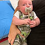 baby, child, pacifier, camouflage_clothing, overalls, infant, person, holding, hand, hospital, medical_equipment, bed, footwear, floor, curious_expression, indoor, adult, skin, clothing, furniture