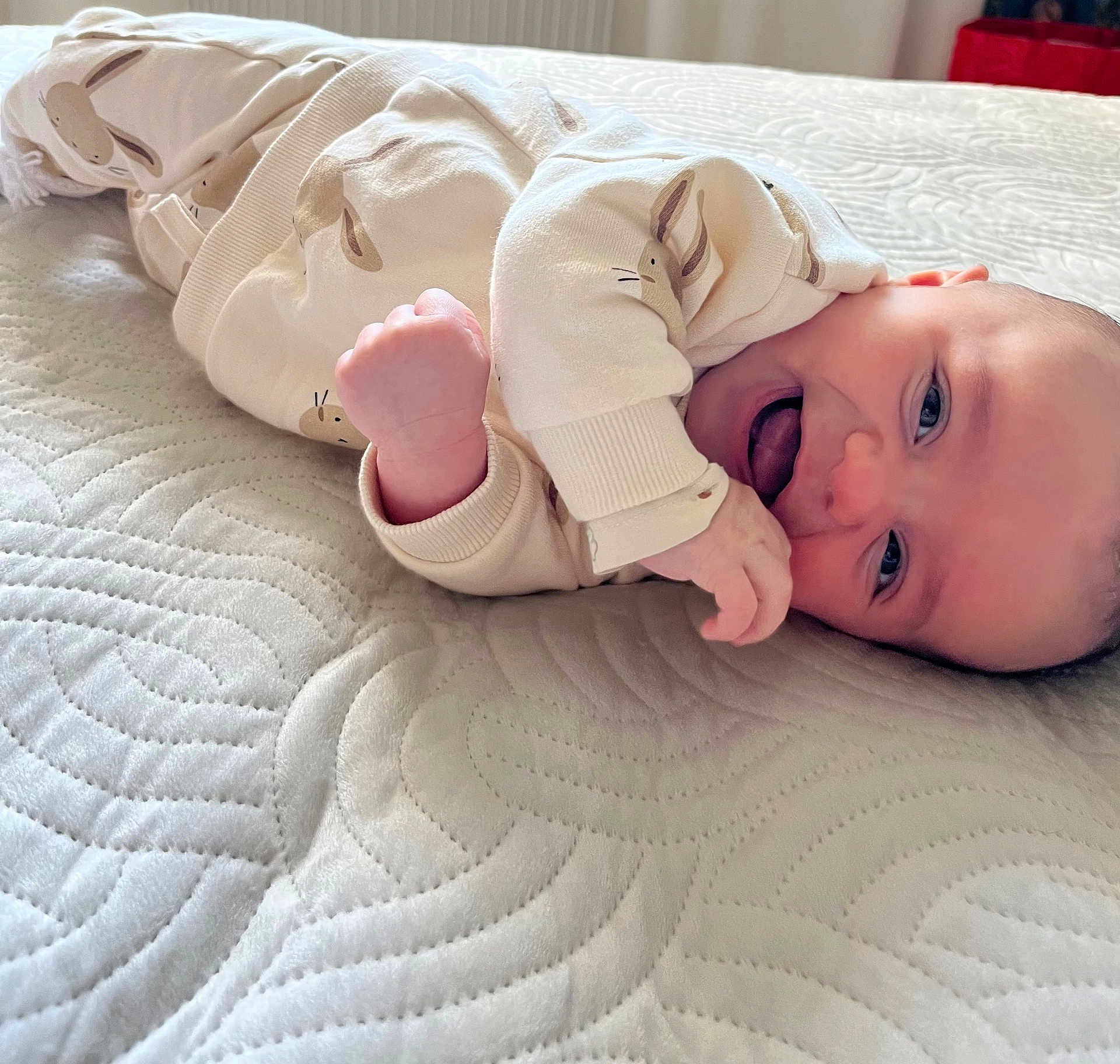 Marius participe au concours pour gagner de l'argent avec cette photo : baby, infant, smiling, tongue_out, lying_down, onesie, bunny_print, bed, quilt, texture, cute, child, happy, skin, face, hand, cozy, indoors, person, portrait