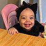 black, bow, child, clothing, curly_hair, cute, earrings, face, hand, happy, headband, high_chair, indoor, person, pink, portrait, smiling, table, toddler, wood