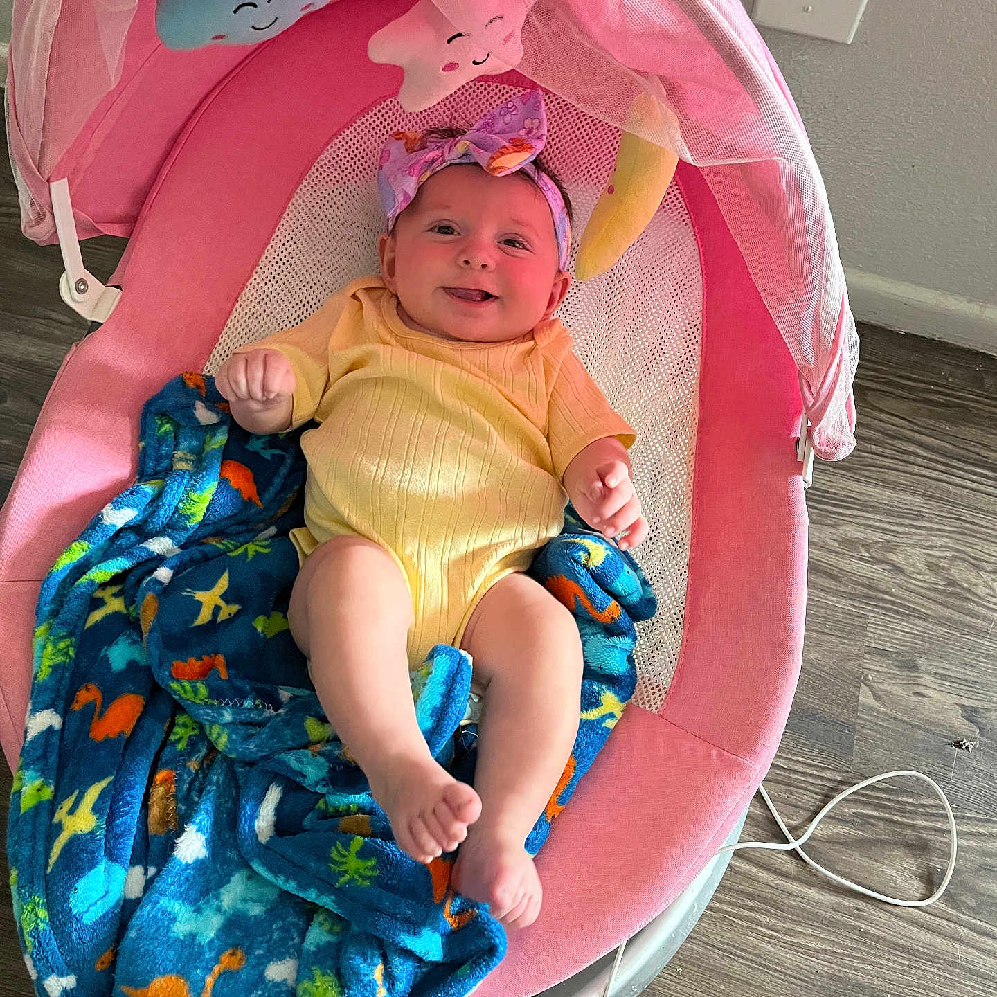 Anluisa is registered to the contest to win money with this photo: baby, infant, pink_baby_rocker, yellow_onesie, headband, blue_blanket, dinosaur_pattern, happy, smiling, indoor, wooden_floor, baby_toy, baby_accessory, child, person, cozy, relaxed, cute, playful, resting