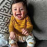 Santi is registered to the contest to win money with this photo: baby, child, smiling, sitting, couch, pillow, clothing, striped_shirt, patterned_pants, white_socks, indoor, happy, cute, portrait, face, hand, infant, home, fabric, cozy