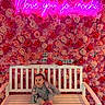 baby, bench, flowers, neon_sign, pink, red, white_bench, child, smiling, indoor, portrait, cute, happy, cozy, colorful, wall_decor, floral_wall, neon_light, casual_clothing, socks