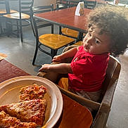 Roman joined the competition — help win amazing prizes! child, toddler, curly_hair, high_chair, pizza, plate, restaurant, table, chair, food, indoor, person, casual_clothing, red_shirt, yellow_shorts, window, daylight, floor, expression, seated