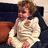 toddler, child, curly_hair, white_outfit, sitting, couch, blue_cushion, brown_cushion, indoor, portrait, cute, young_child, clothing, long_sleeve, pants, home, furniture, relaxed, person, expression