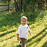 toddler, child, grass, outdoor, sunlight, trees, nature, walking, casual_clothing, white_shirt, pants, sneakers, greenery, daylight, forest, shadows, young_child, curious, person, park